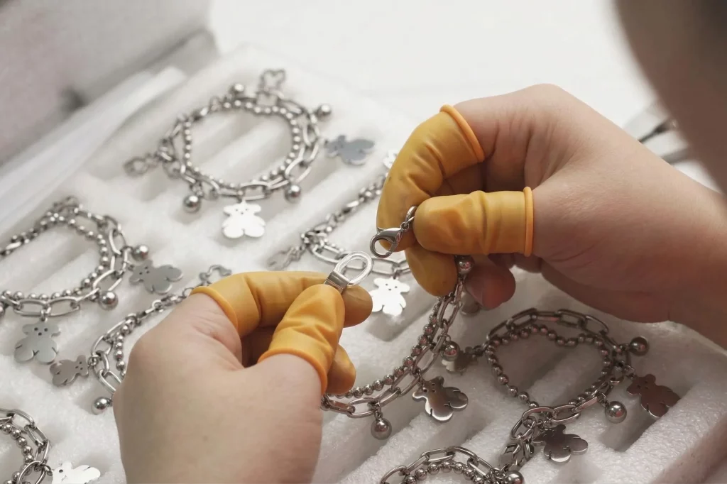 Pendant bracelet inspection – hands with finger cots, stainless steel charm bracelet on beaded chain, bulk order samples in tray, wholesale jewelry quality assurance