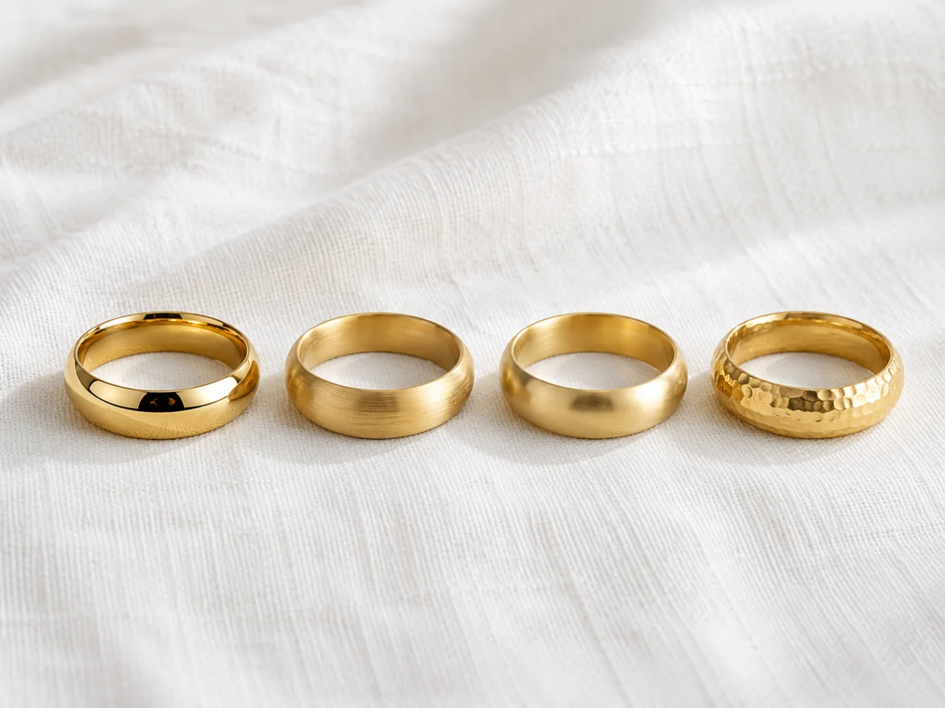 Four different gold rings arranged horizontally on a white fabric background, showcasing PVD coating, gold plating, gold-filled, and solid gold finishes.
